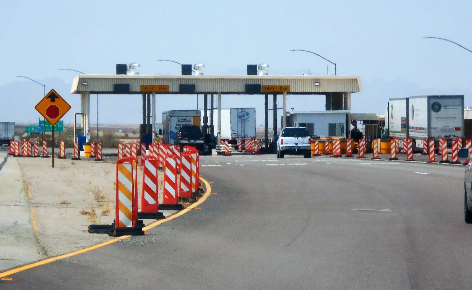 Commercial vehicles approaching a border inspection checkpoint with traffic cones and inspection booths on a highway